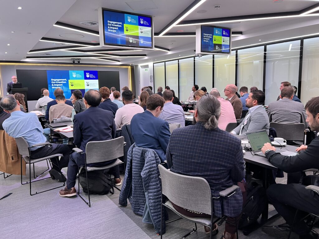 Clean Maritime Research Partnership members looking at a presentation introducing the Clean Maritime Research Hub subgroups, hosted by Tony Roskilly, director of the UK National Clean Maritime Research Hub. Attendees are seated around tables, the room is full. 